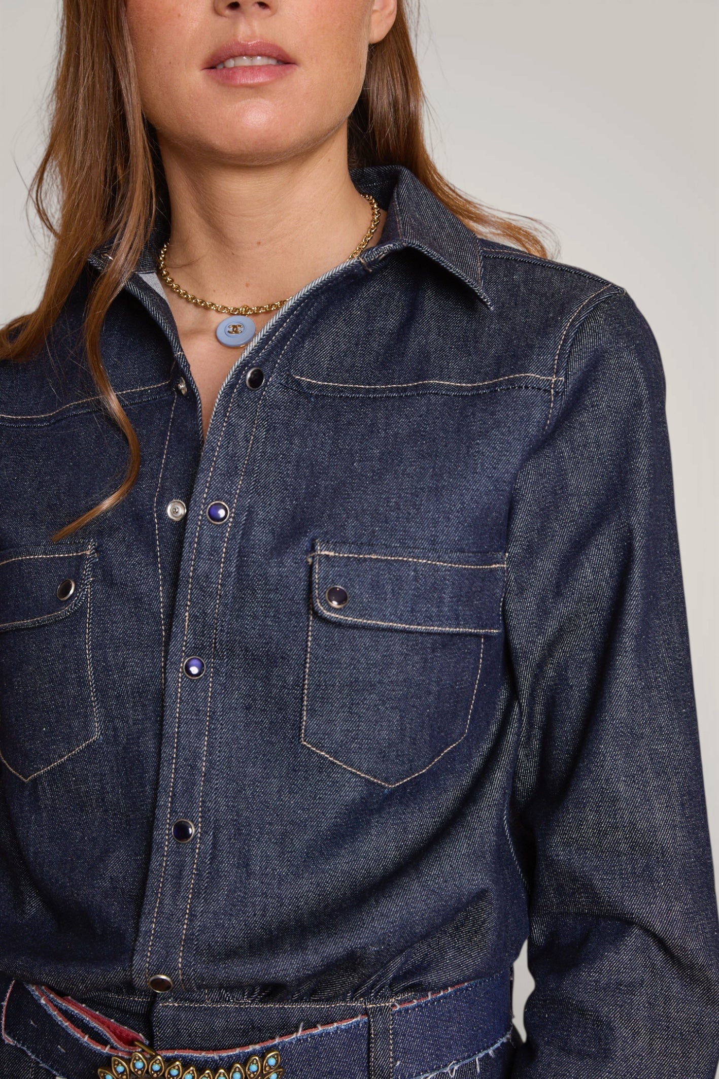 Gros plan sur la chemise en jean de la combinaison et ses boutons pressions en nacre indigo. Ceinture en denim portée avec.